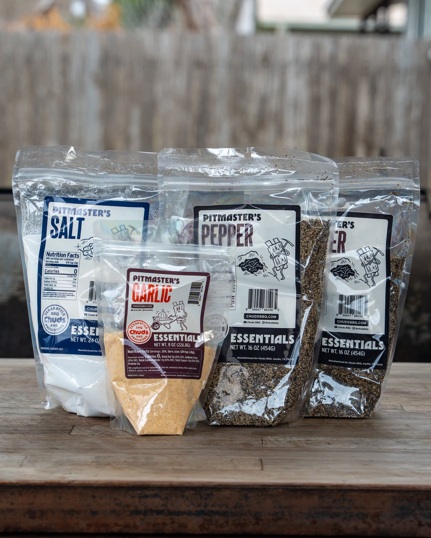 Three plastic bags labeled Pitmaster's Salt, Pitmaster's Pepper, and Pitmaster's Garlic, placed on a wooden surface.